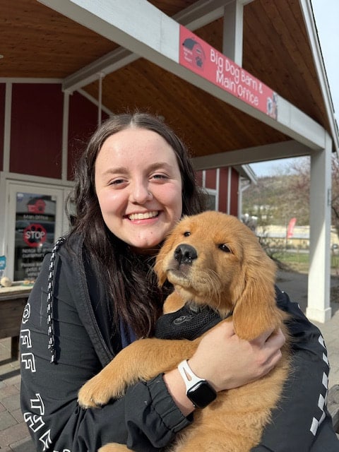 A smiling staff member hugging a golden retriever puppy under the Big Dog Barn sign